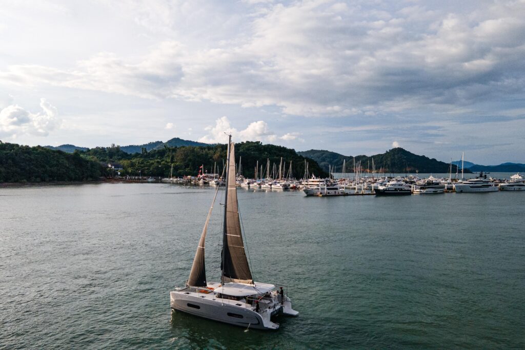 Sailboat "Excess 11" 2025 model cruising near a marina with lush hills in the background.