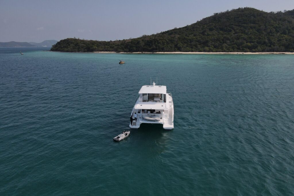 White catamaran anchored in turquoise water near a tropical island. Dinghy alongside.